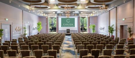 Un salón de conferencias con filas de sillas, un escenario con un logotipo y un amplio ventanal con vistas a zonas verdes.