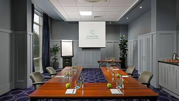 Una sala de conferencias con una larga mesa, sillas, una pantalla de proyector y una planta.