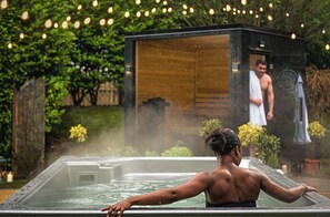 Una bañera de hidromasaje con una persona relajándose, rodeada de vegetación e iluminada con luces colgantes.