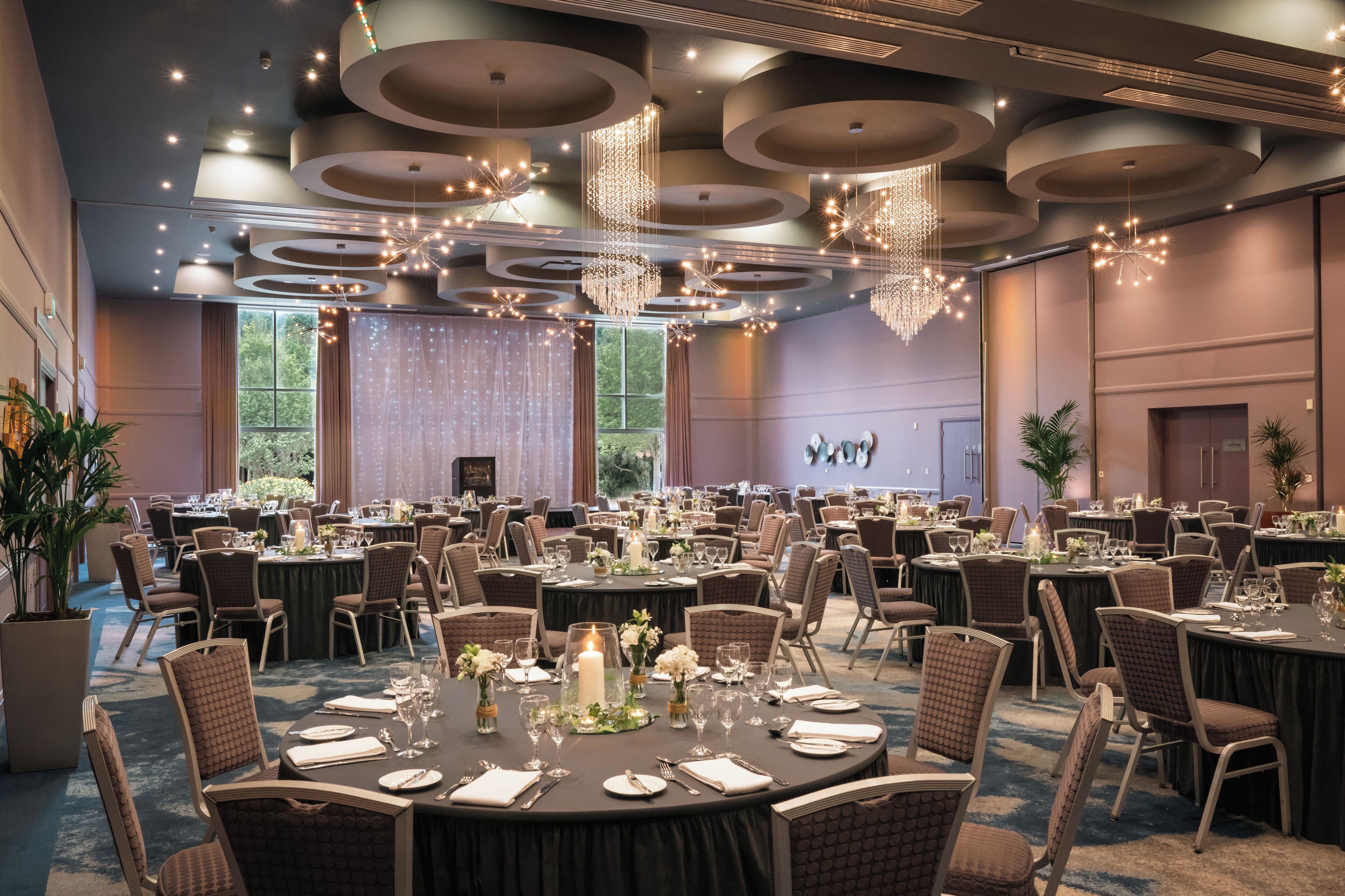A banquet hall with round tables, chairs, chandeliers, and large windows.