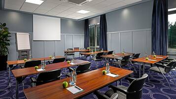Un salón de conferencias con varias mesas, sillas, una pantalla de proyector y un ventanal con cortinas.