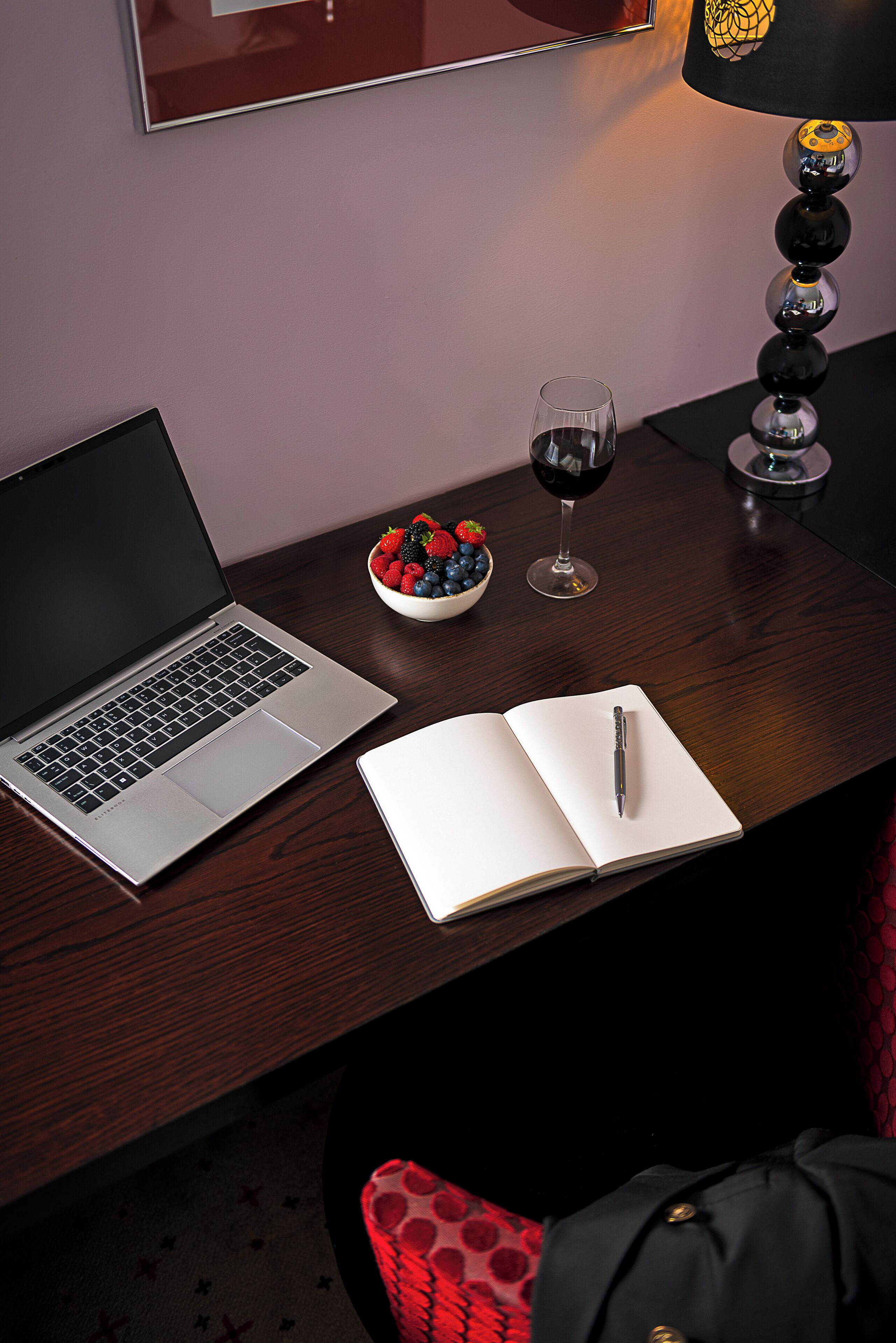 A laptop, notebook, pen, bowl of fruit, wine glass, and a lamp on a wooden desk.