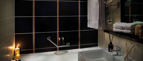 A modern bathroom with a large bathtub, a separate sink, and a mirror with a light. There are candles, a towel, and toiletries on the counter.