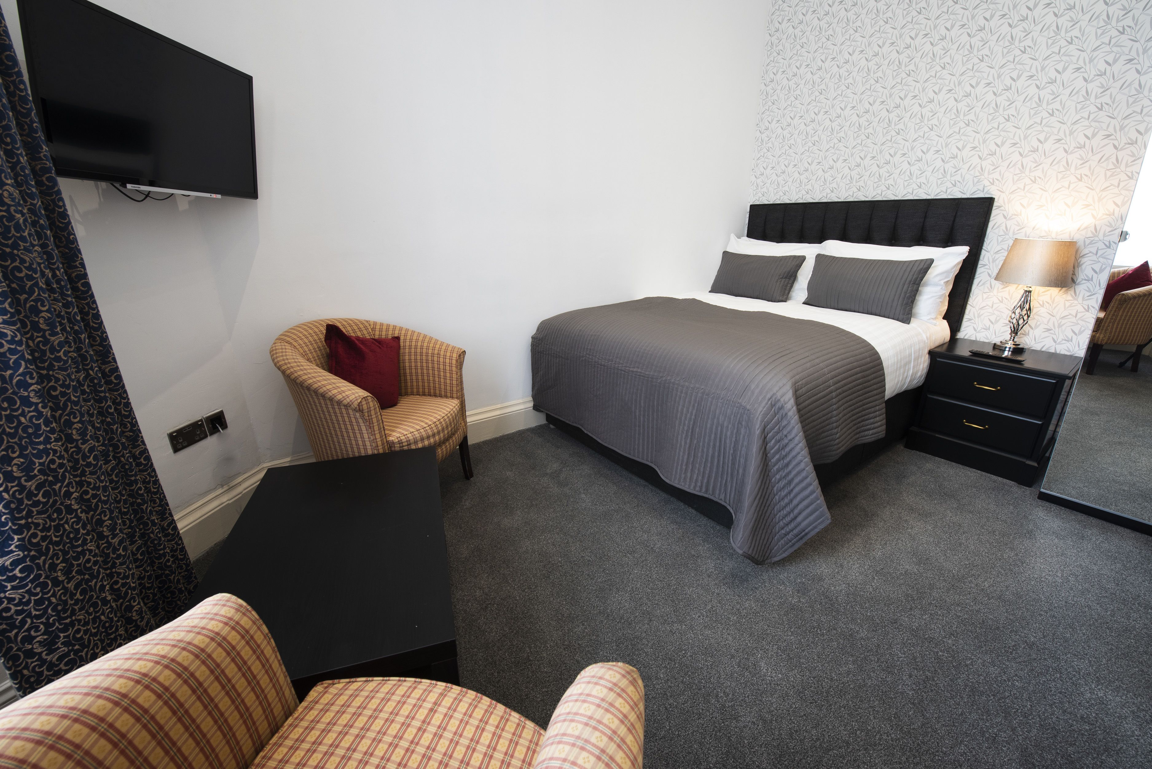 Ein Hotelzimmer mit Bett, Sessel, Schreibtisch, an der Wand montiertem Fernseher und Nachttisch mit Lampe.