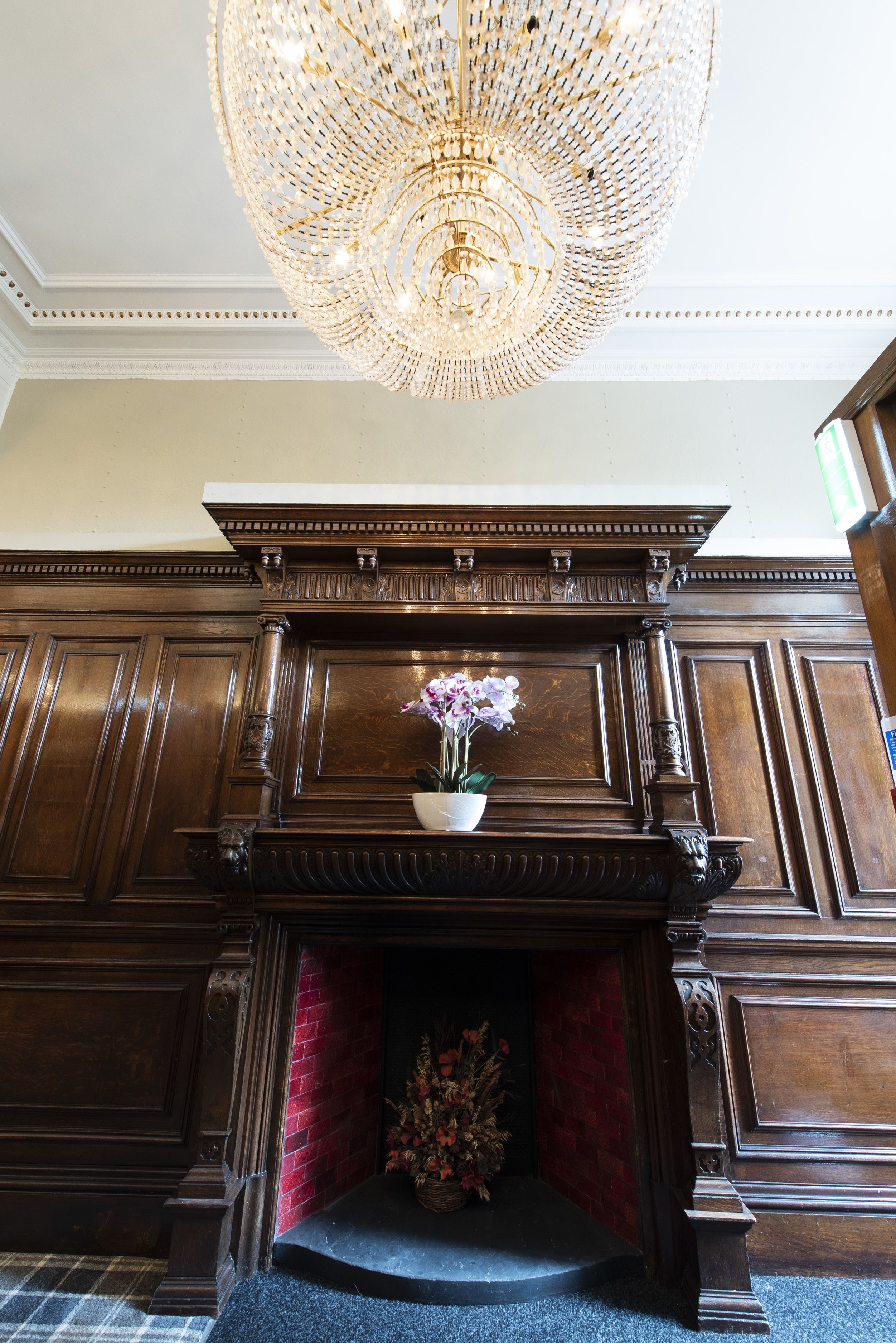 A grand room with a large, ornate chandelier, a wooden mantelpiece, and a fireplace with a red brick surround.
