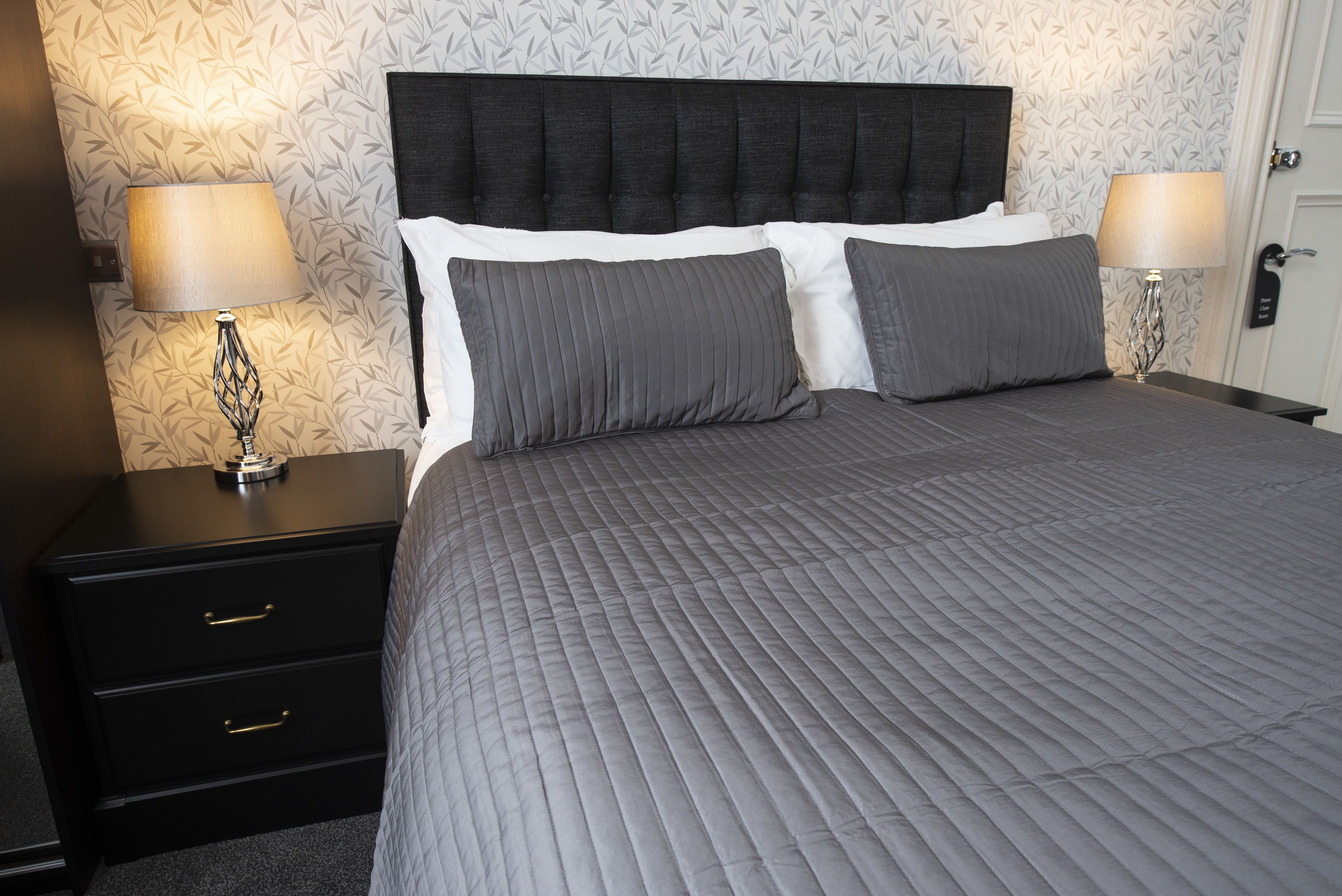 Una cama bien tendida con un cabecero oscuro y ropa de cama gris, flanqueada por dos mesitas de noche con lámparas y un cajón pequeño.