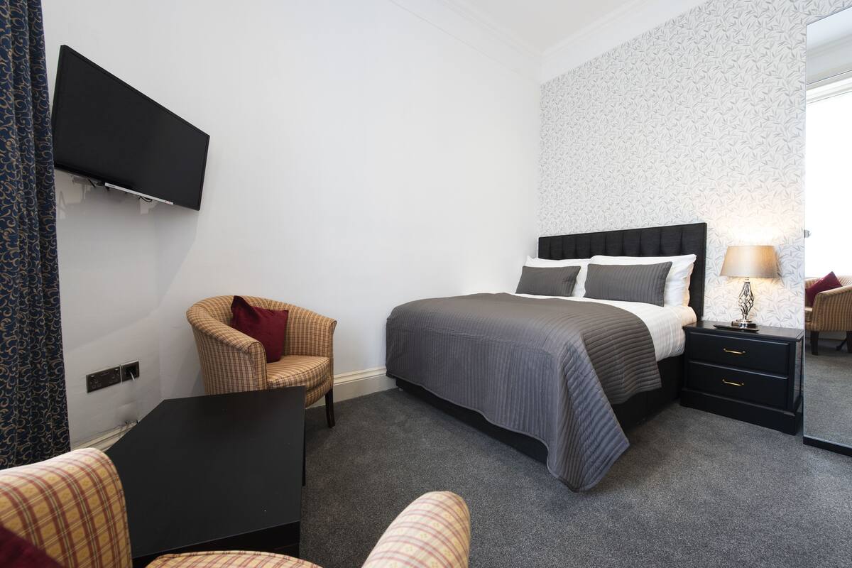 A hotel room with a bed, a chair, a desk, a TV mounted on the wall, and a nightstand with a lamp.