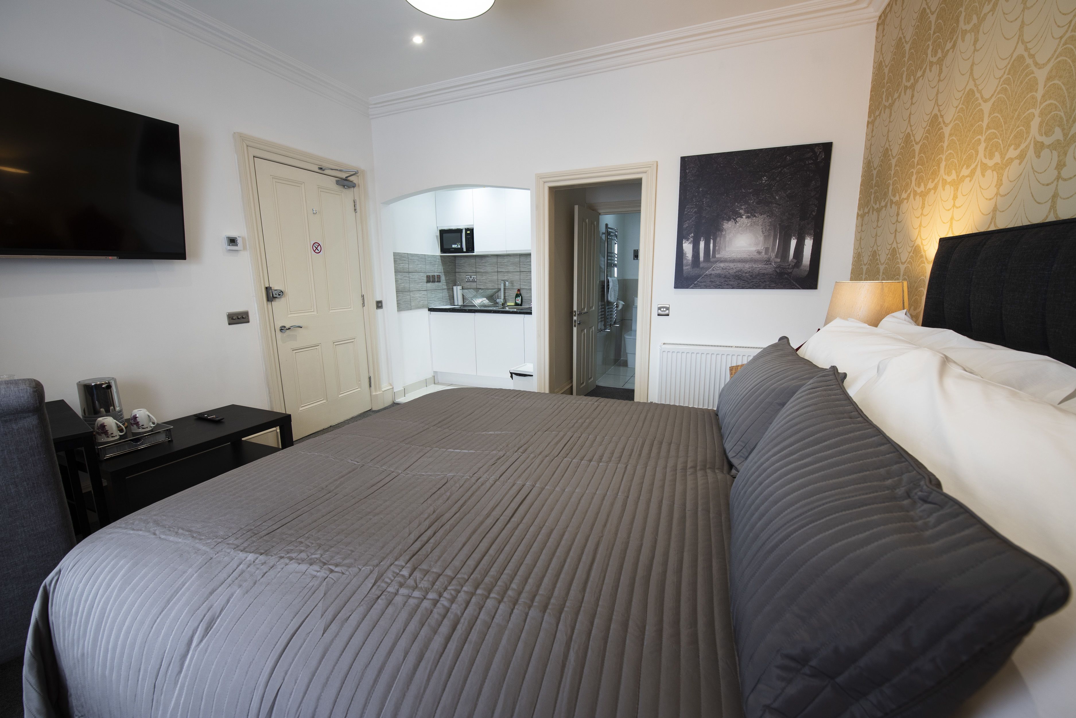 A bedroom with a large bed, a TV mounted on the wall, a desk with a chair, and a door leading to a bathroom.