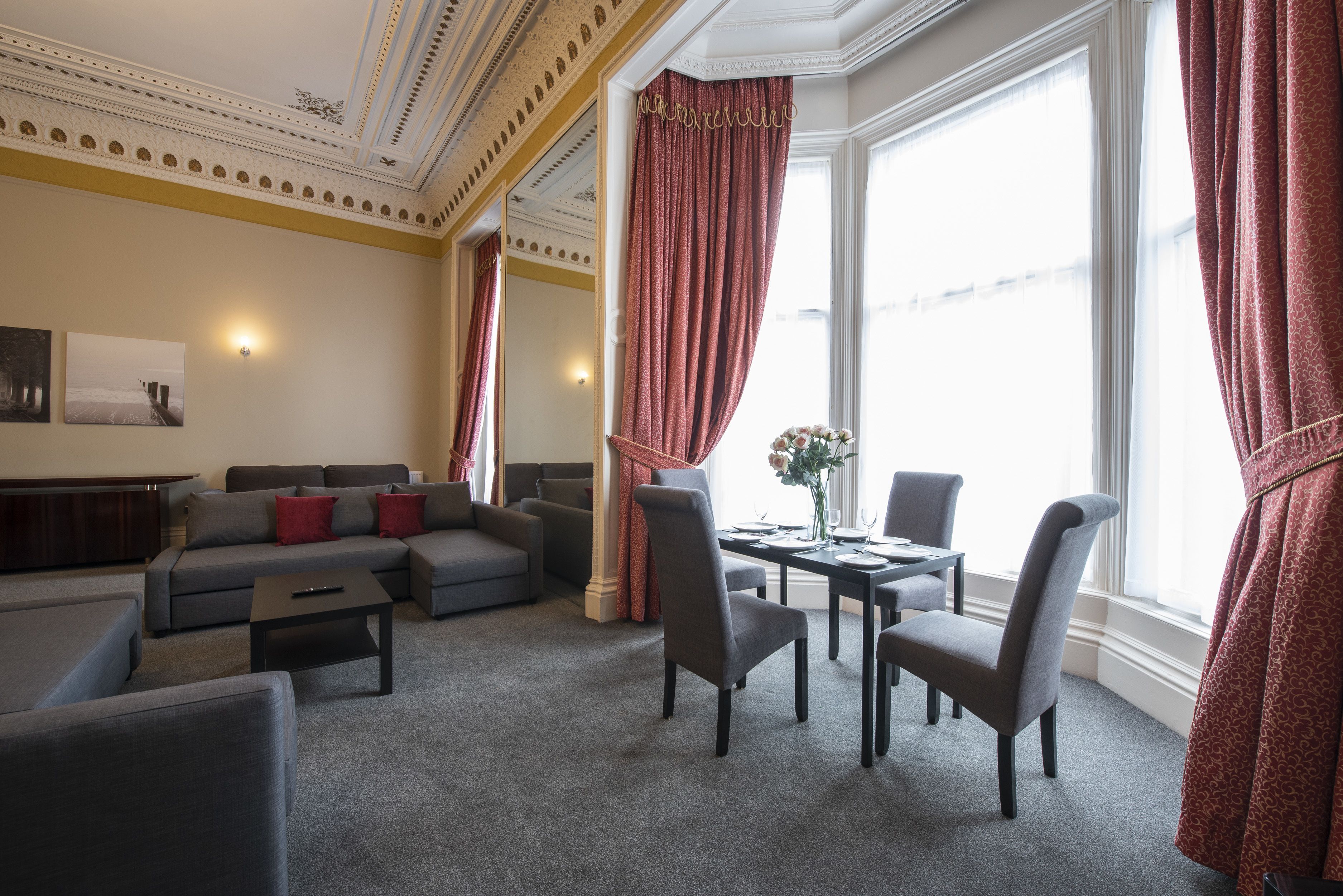 A spacious living room with a gray sofa set, a dining table with chairs, and a large window with red curtains.