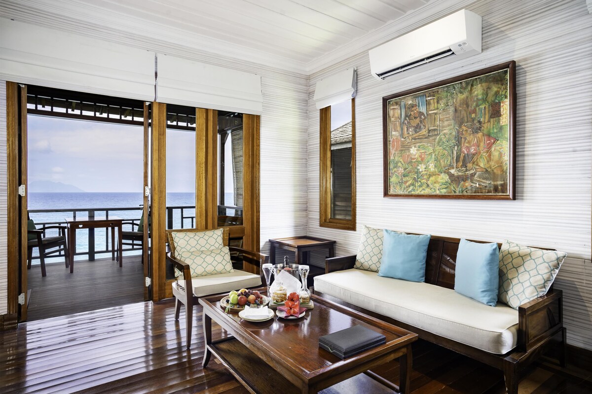 A room with a wooden floor, a sofa with patterned cushions, a dining table with fruit and a bottle, and a painting on the wall.