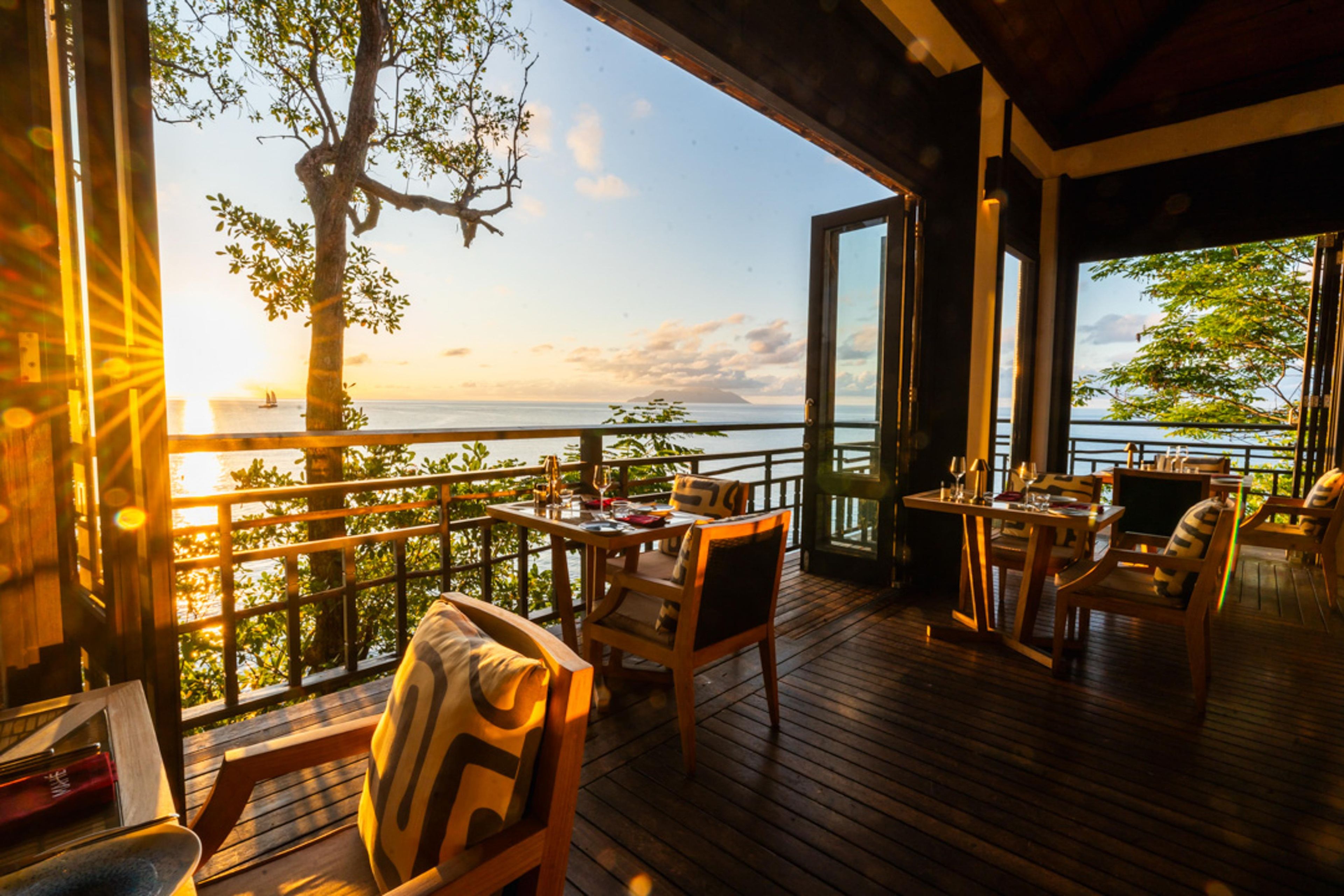 An outdoor deck with wooden flooring, furnished with chairs and tables, overlooking a body of water and a tree.