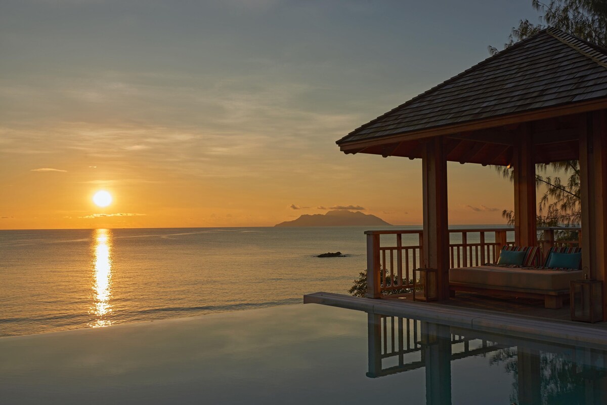 A serene sunset view from an infinity pool overlooking the ocean, with a wooden gazebo and lounge area.