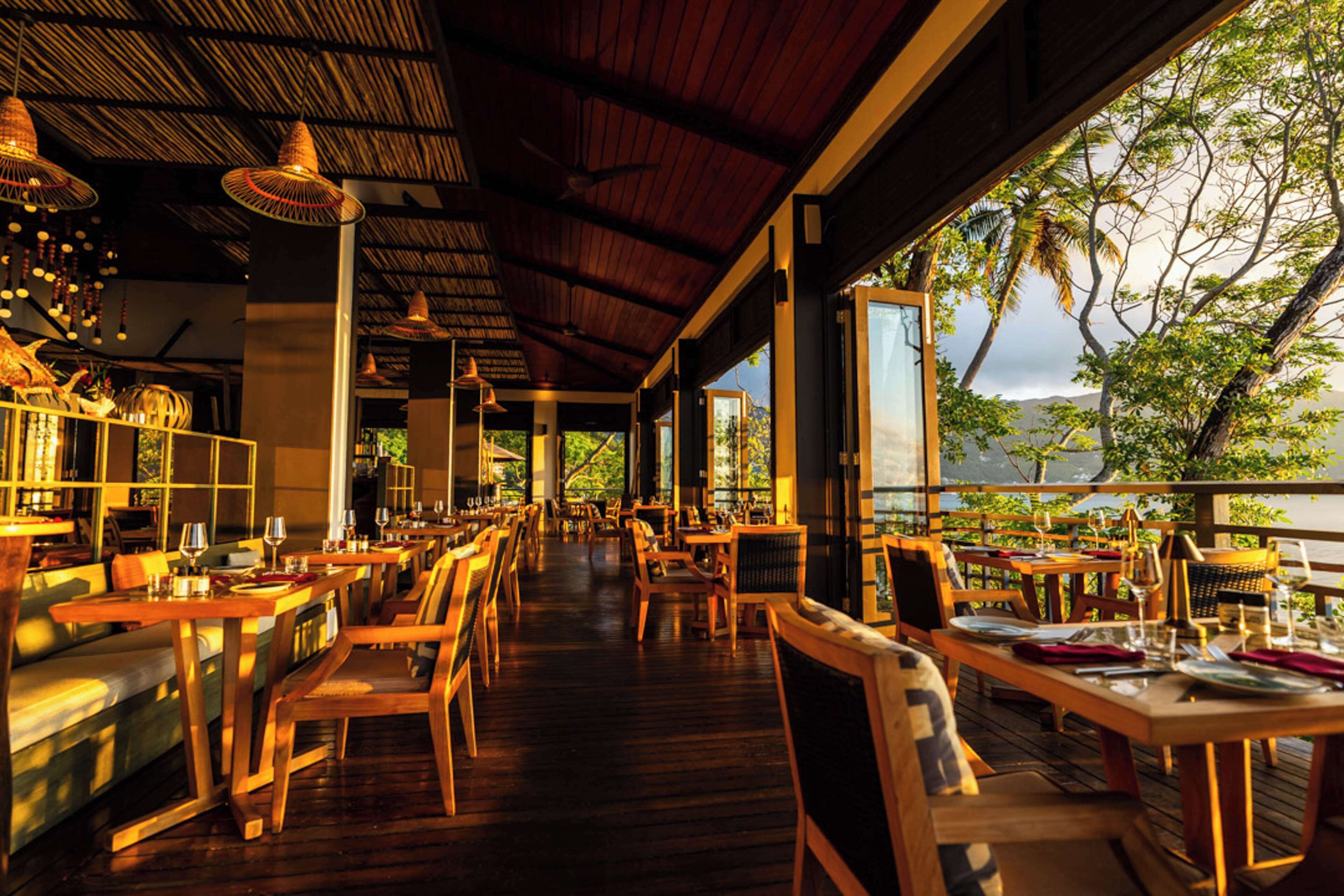 An indoor dining area with wooden tables and chairs, large windows, and a view of the outdoors.