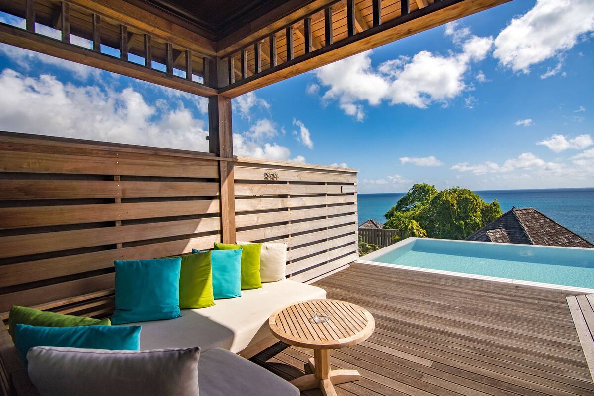Eine Holzterrasse mit Pool, ein weißes Sofa mit bunten Kissen und Blick aufs Meer.