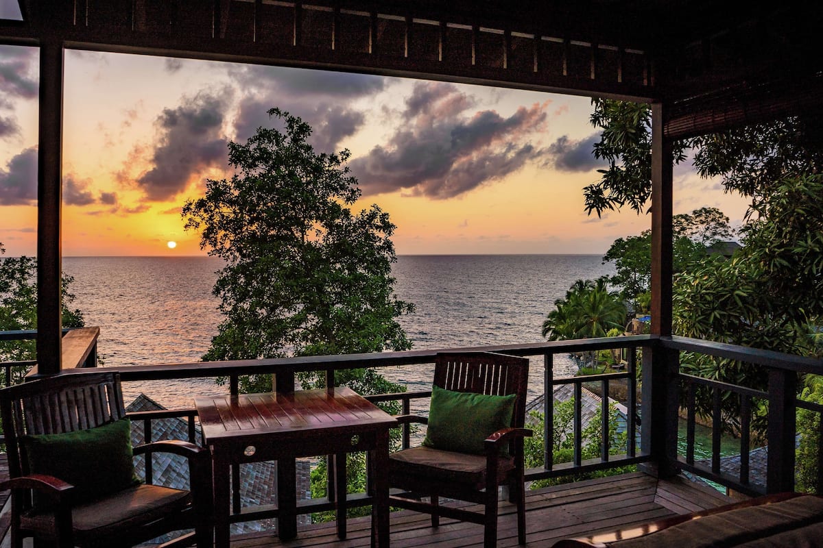 Eine Holzterrasse mit Tisch und Stühlen, mit Blick auf einen Sonnenuntergang über dem Meer.