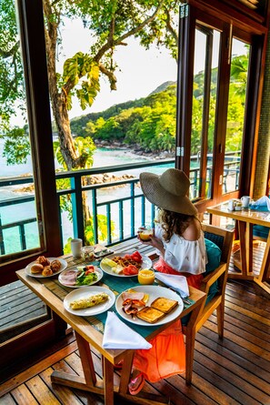 A dining area with a table set for two, overlooking a scenic view of a beach and hills. - Hilton Seychelles Northolme Resort & Spa (Mahé Island)