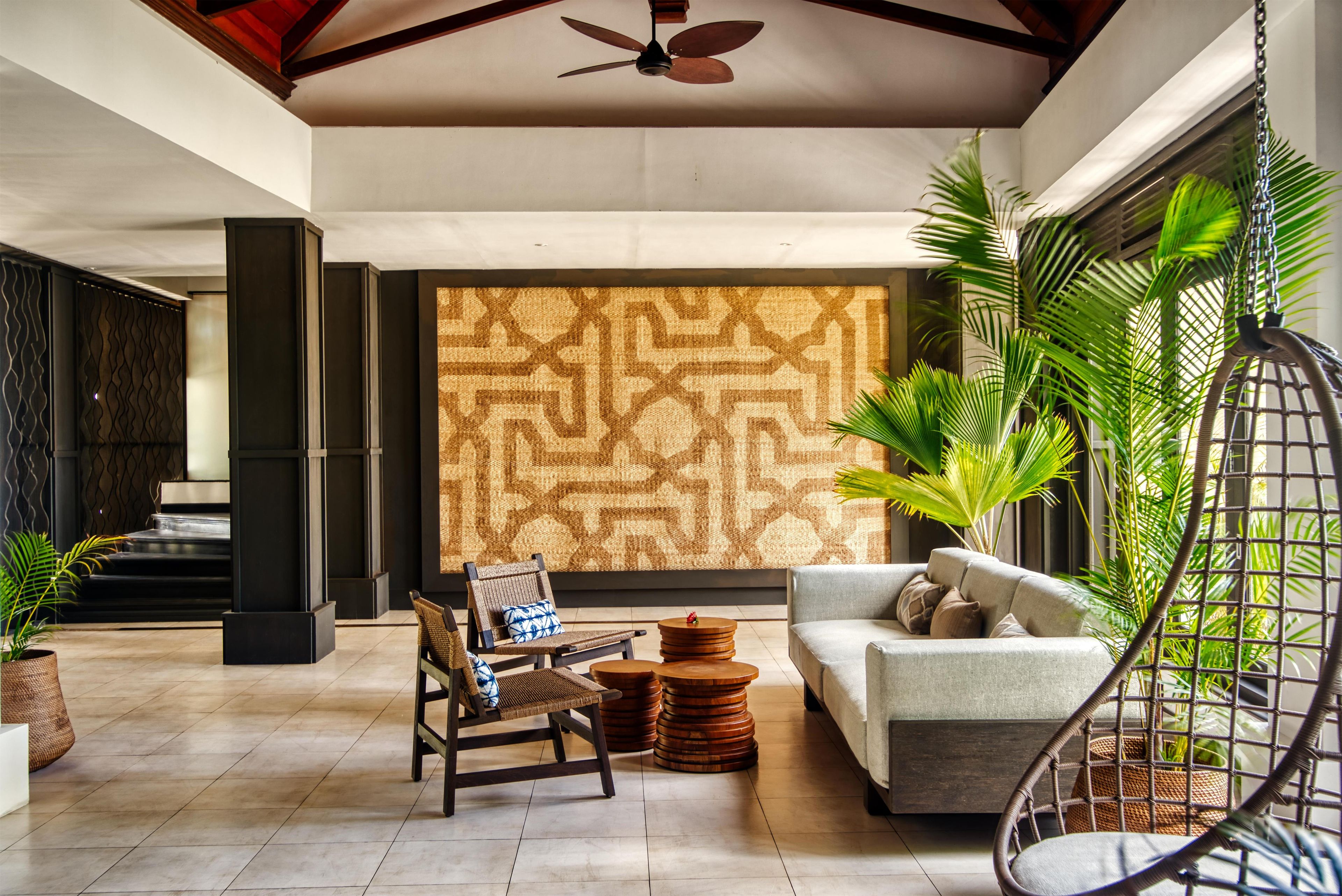 A spacious lobby with a high ceiling, a ceiling fan, a patterned wall, a sofa, a chair, and several potted plants.