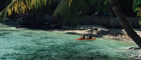Mar turquesa cristalina con un kayak a lo lejos, una playa de arena y un muro de piedra con sillas de descanso.