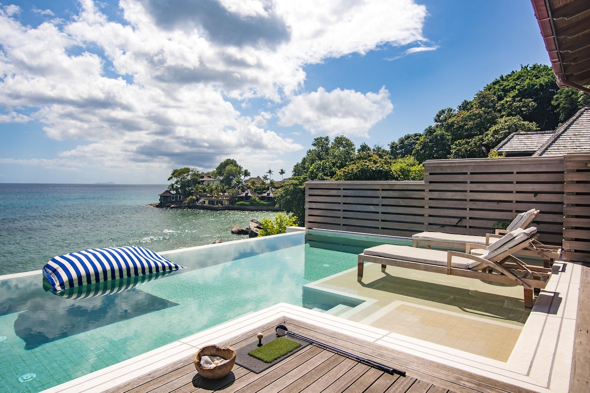 A clear blue infinity pool with lounge chairs and a wooden deck overlooking the sea.