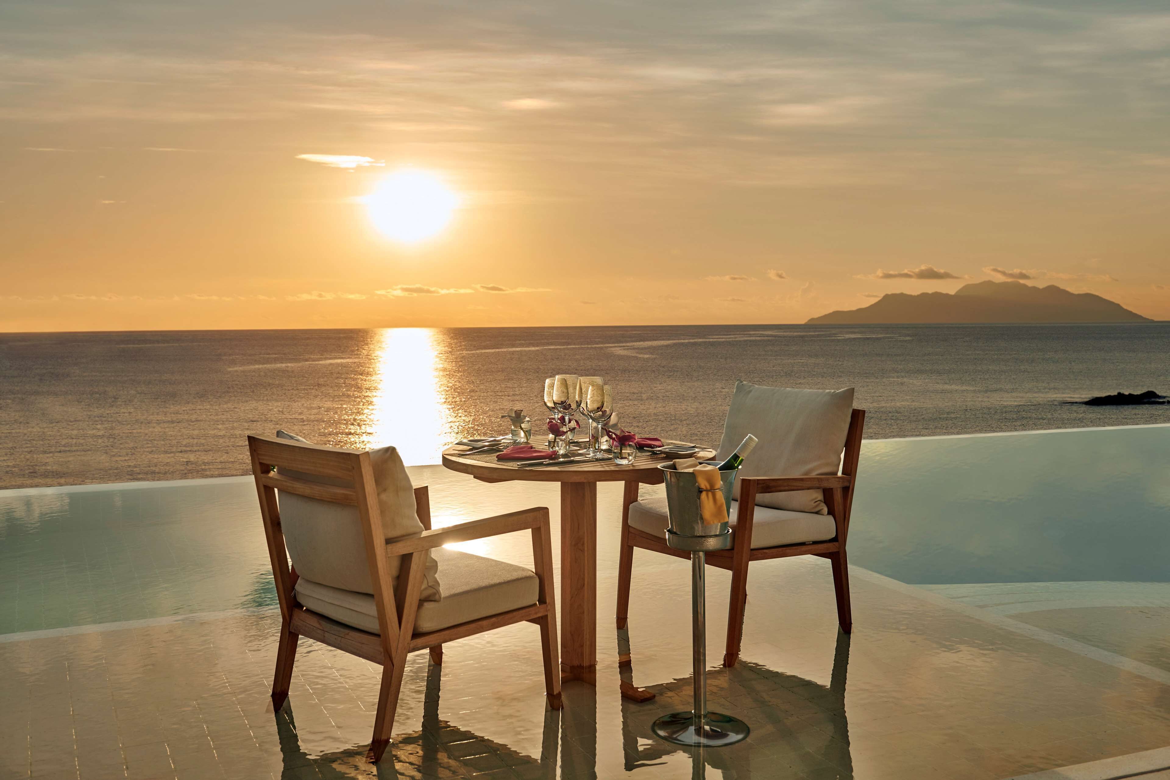 A table with chairs set up by an infinity pool overlooking the sea at sunset.