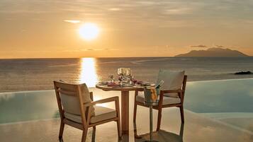 A table with chairs set up by an infinity pool overlooking the sea at sunset.