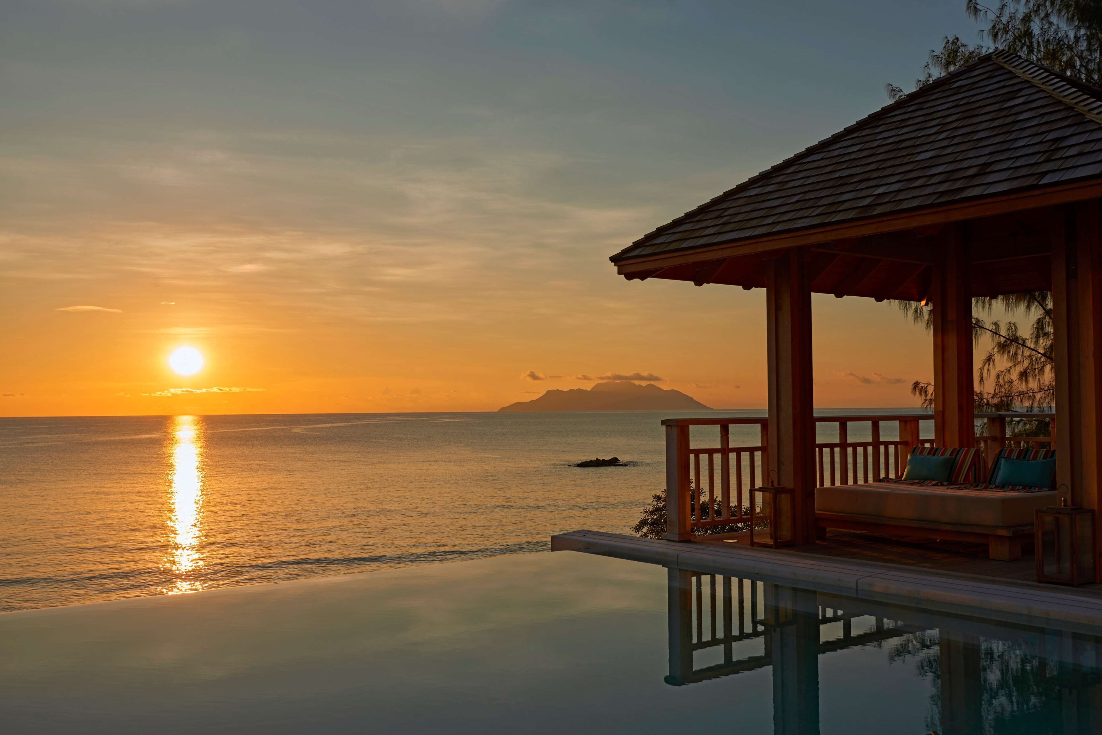 A sunset view from an infinity pool with a wooden deck and seating area overlooking the ocean.