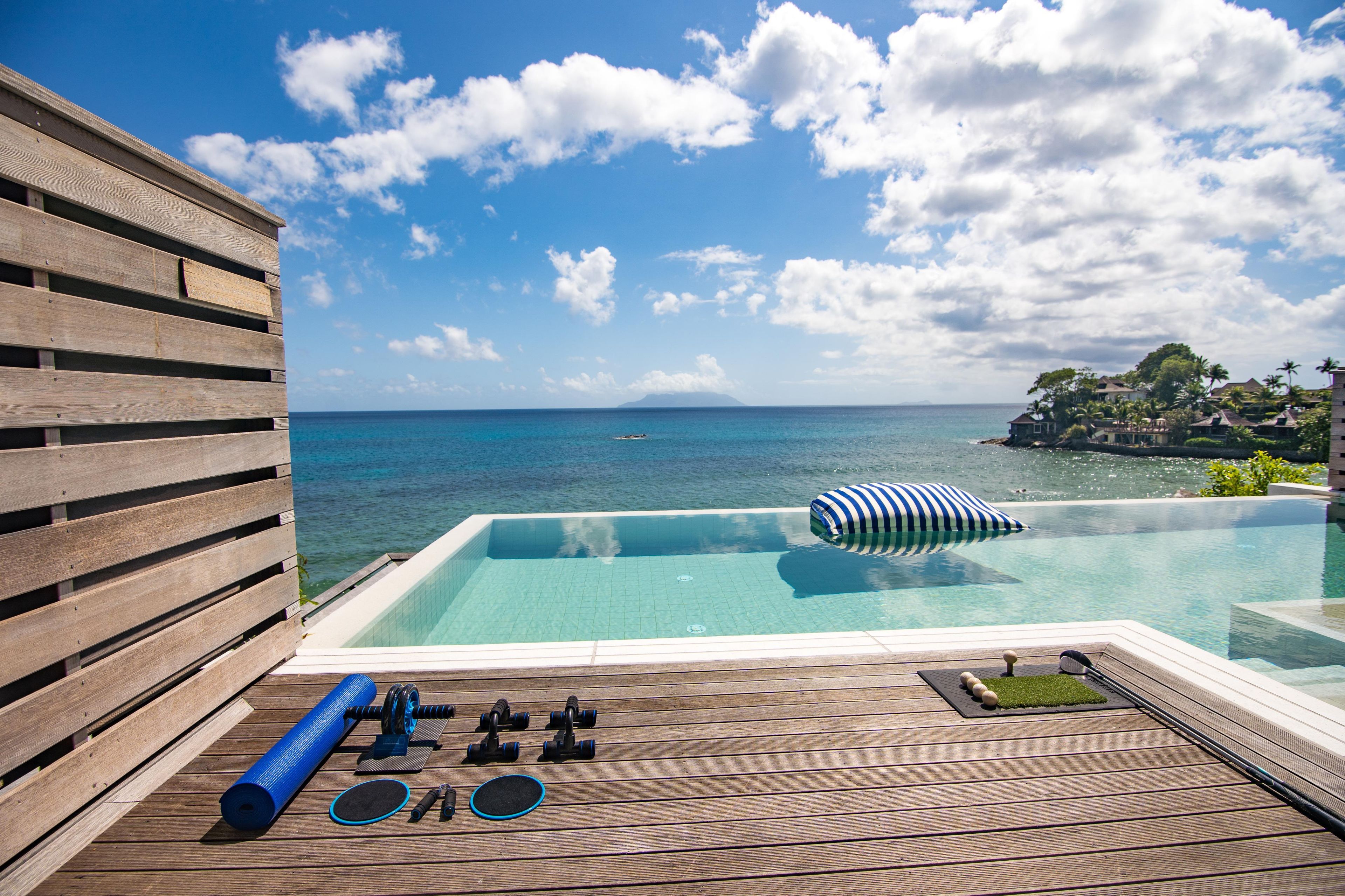 A clear blue sky, a wooden deck, a pool with a view of the sea, and a tropical landscape in the background.