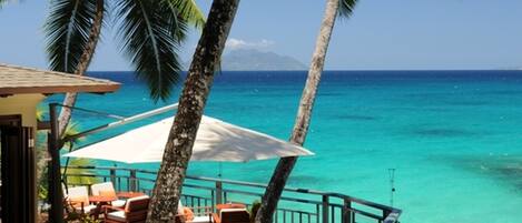 A wooden deck with outdoor furniture overlooking turquoise waters, palm trees, and a clear sky.