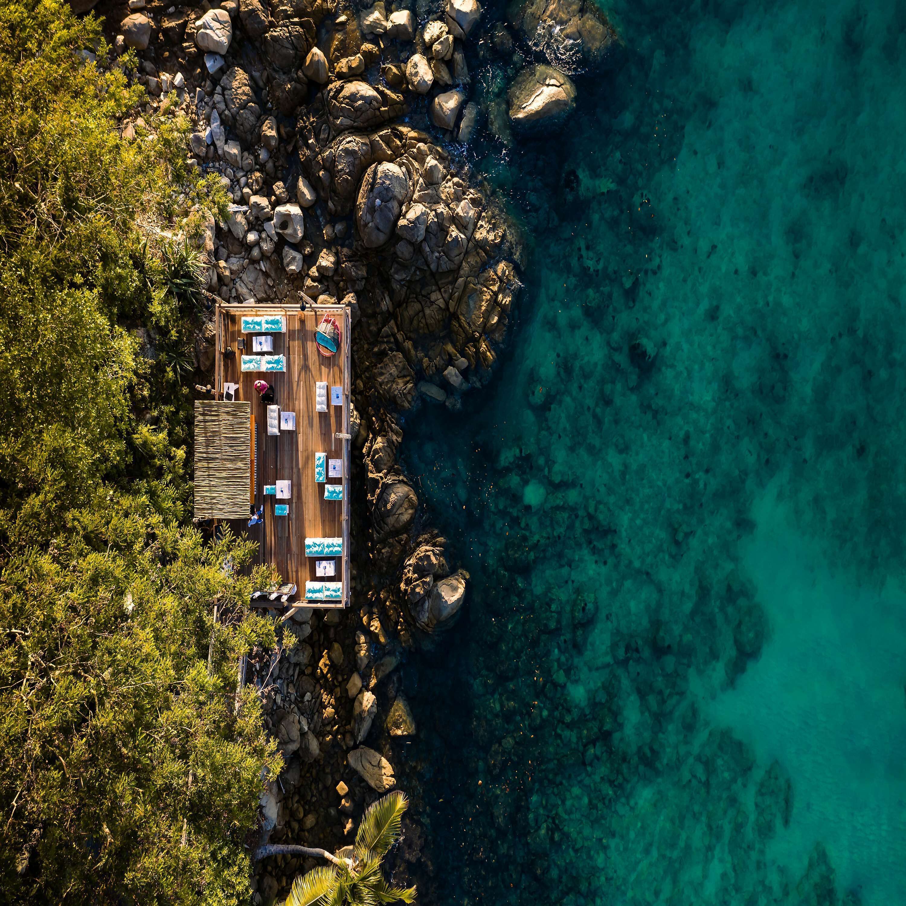 Vue aérienne d’une terrasse en bois dotée de plusieurs pièces, entourée d’un paysage rocheux et bordée d’une eau turquoise cristalline.