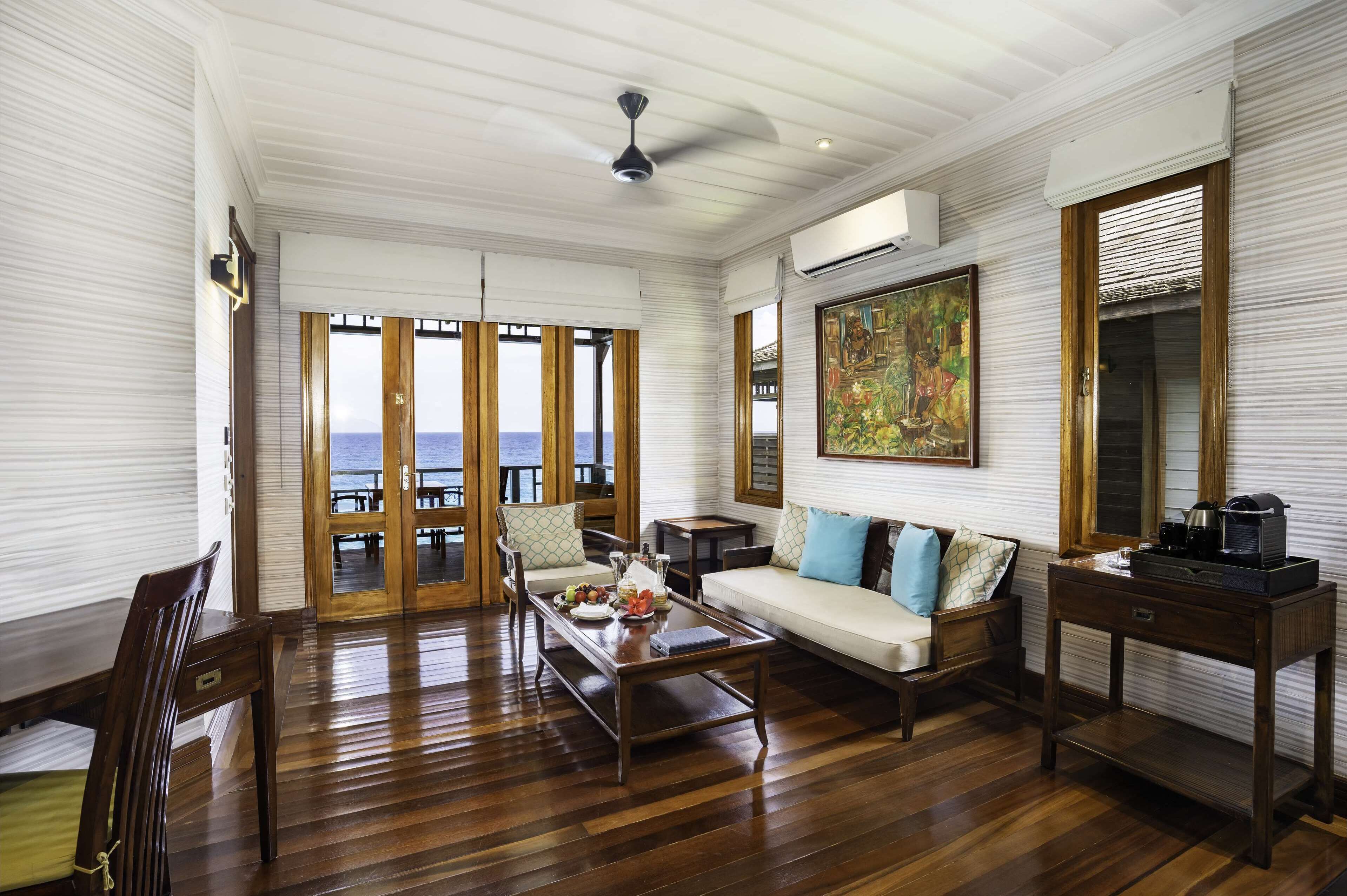 A spacious living room with a wooden floor, a sofa, a coffee table, and a view of the sea through large glass doors.