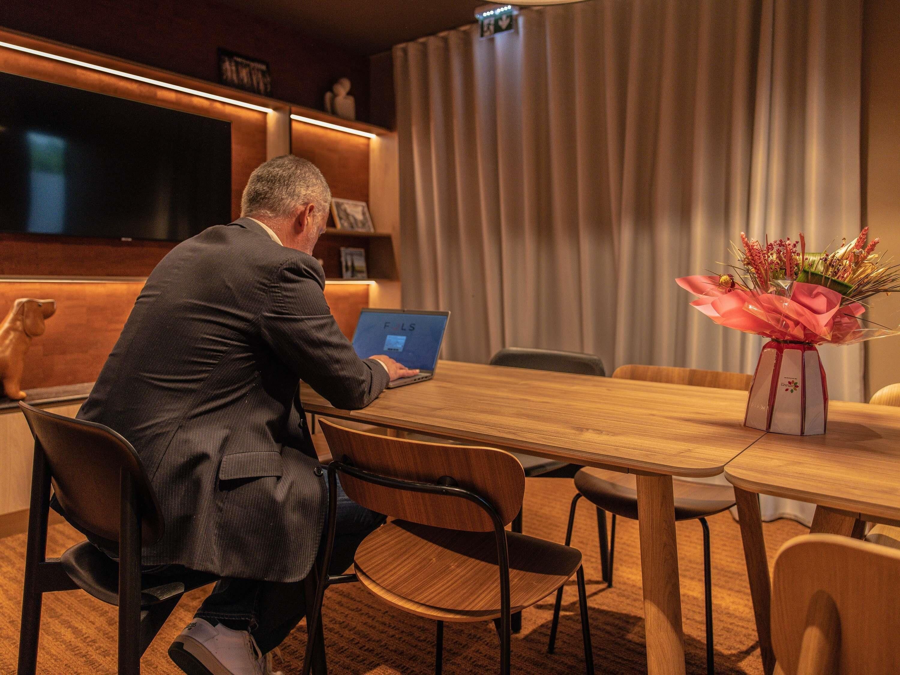 Un hombre sentado en una mesa de madera usando una computadora portátil en una habitación con un ventanal grande, una cortina y un jarrón de flores.