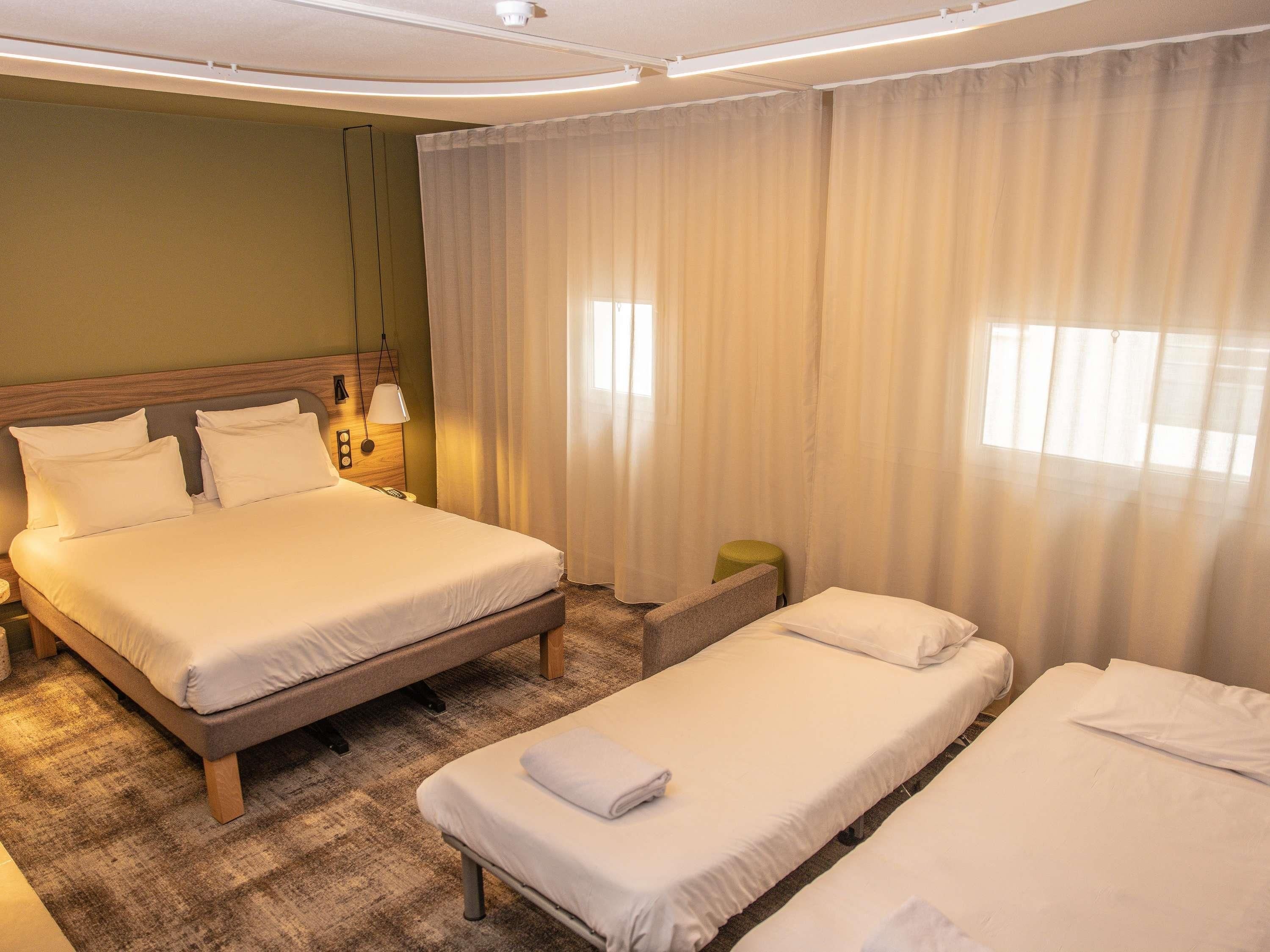 A hotel room with a large bed, a smaller bed, a nightstand, a lamp, and a window with curtains.