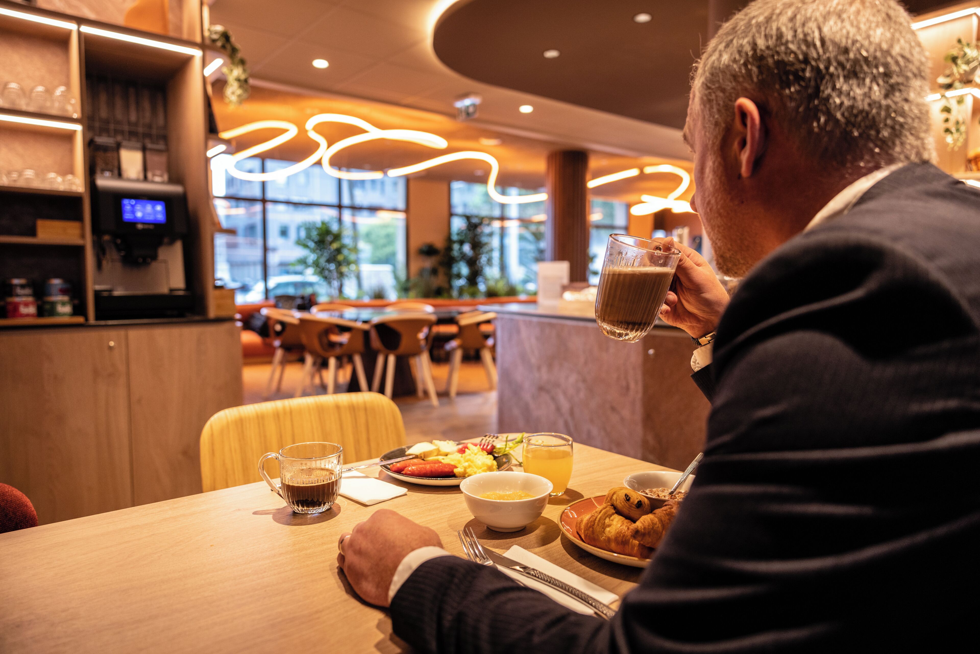 Un homme est assis à une table, devant un repas, une machine à café, et il a une vue sur l’extérieur.