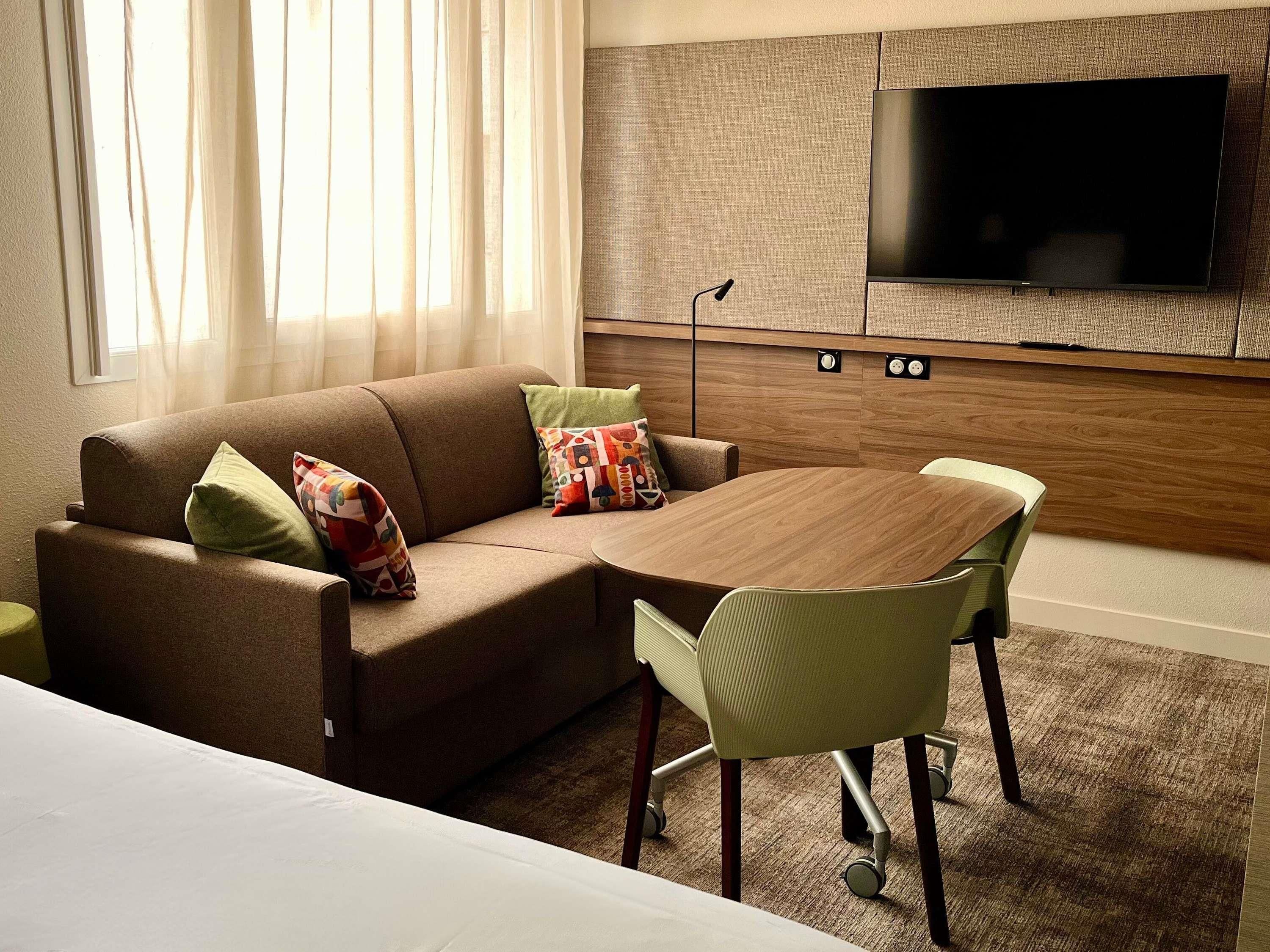 A modern hotel room with a sofa, a dining table, and a TV mounted on the wall.