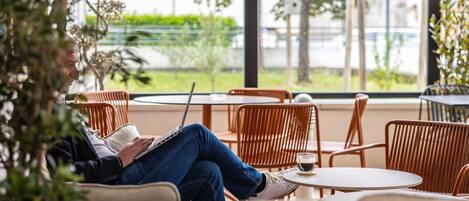 A person sitting on a chair with a laptop, in a room with round tables and chairs, near a window with a view of greenery.