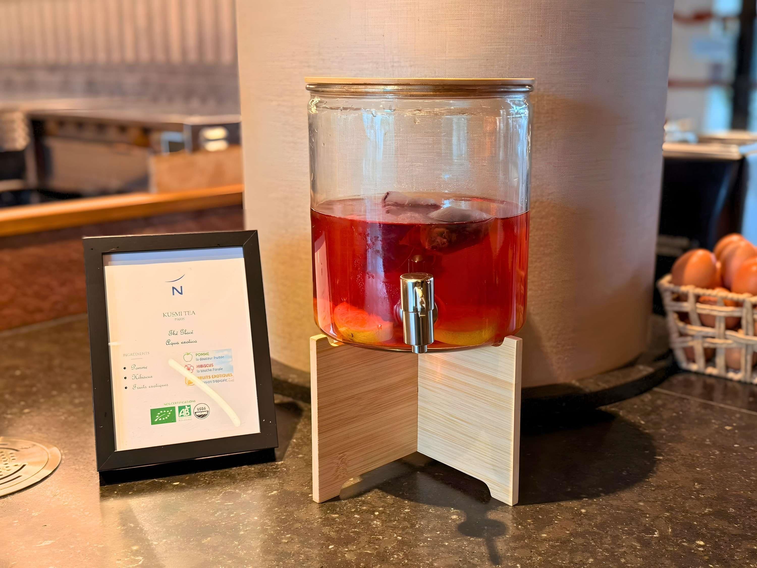 A glass jar with a metal tap filled with a red liquid, placed on a wooden stand.