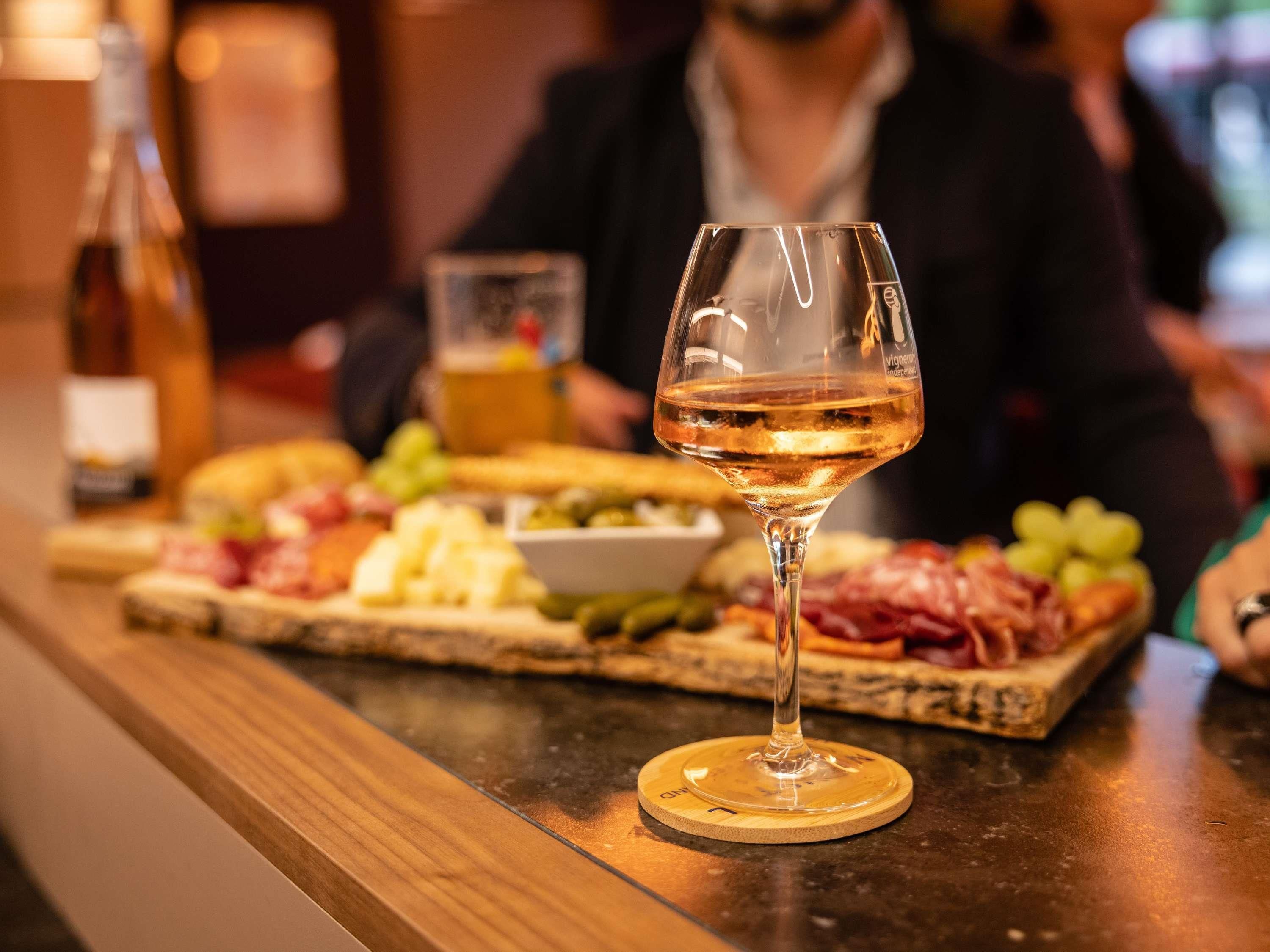 A wine glass on a wooden coaster at a bar with a variety of cheeses, meats, and fruits.