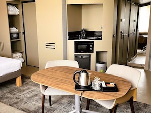 A hotel room with a bed, a wooden table with a kettle, a microwave, and a trash bin.
