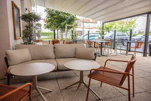 A café with a sofa, round tables, and wicker chairs. - Novotel Suites Clermont Ferrand Polydome (Clermont-Ferrand)