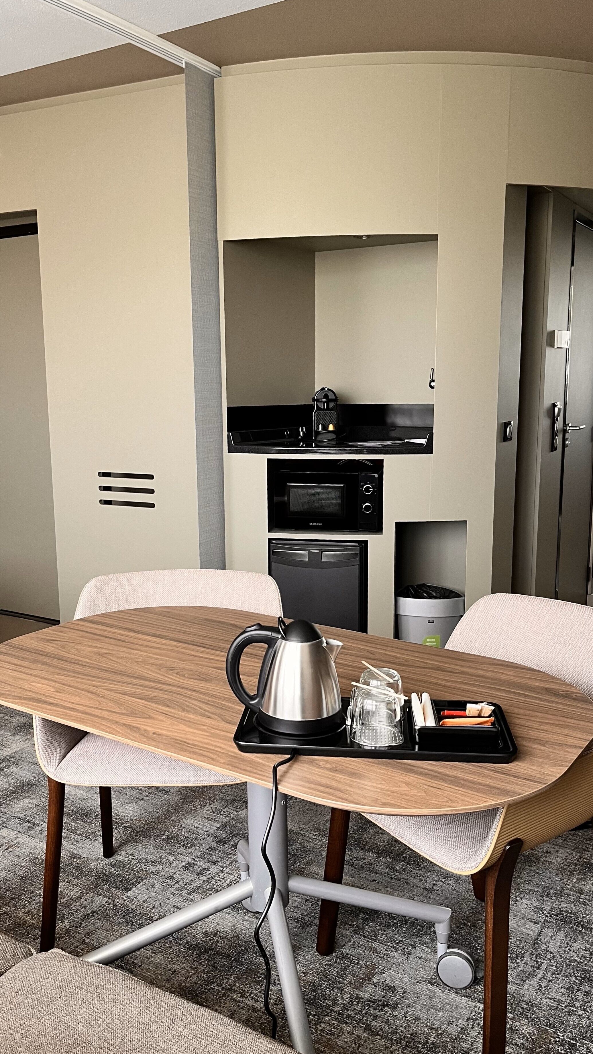 A compact hotel room with a kitchenette, a wooden dining table with a kettle, and a small coffee maker.
