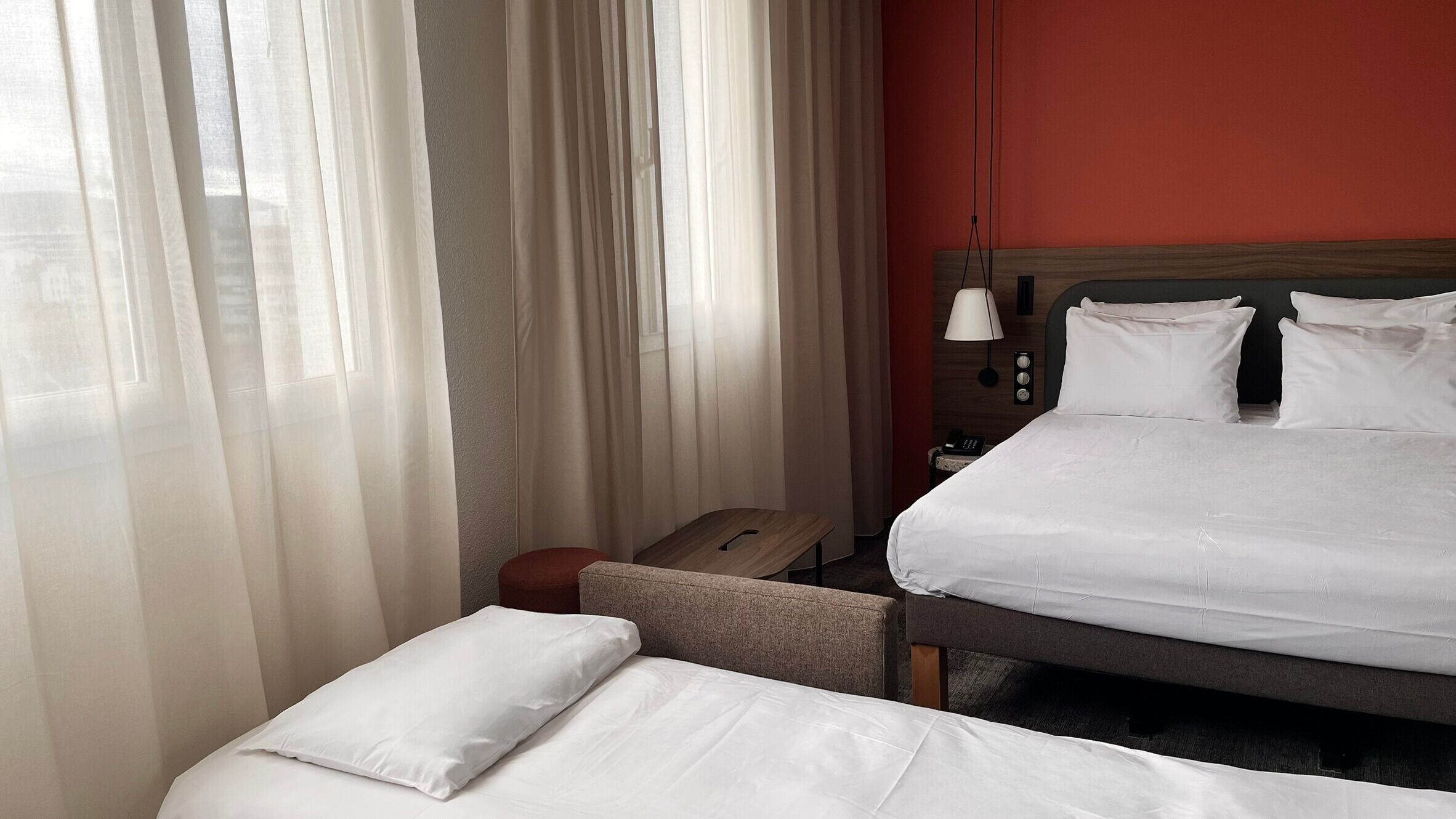 A hotel room with a bed, a bedside table, a lamp, and a window with sheer curtains.