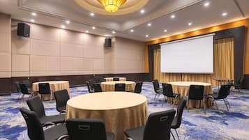 Un salón de conferencias con mesas redondas, sillas negras, una pantalla de proyector y suelo alfombrado.
