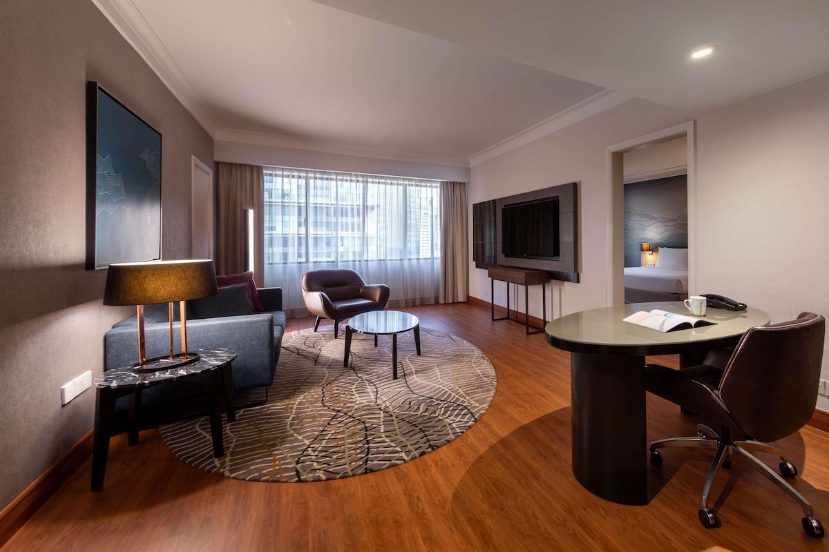 A hotel room with a sofa, armchair, round table, TV, and a bed with a headboard.