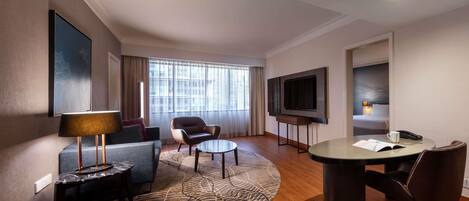 A hotel room with a sofa, armchair, round table, TV, and a bed with a headboard.