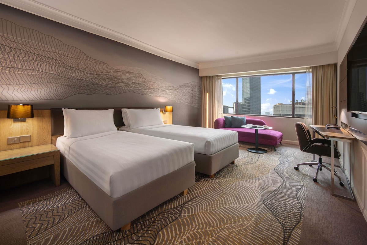 A hotel room with two beds, a desk, a chair, a sofa, and a large window with a city view.