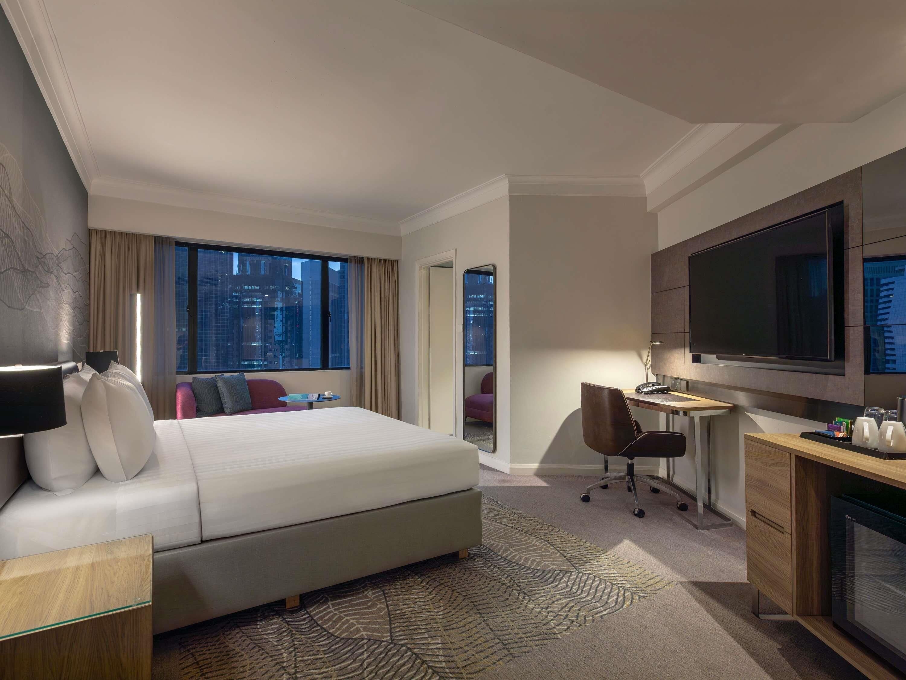 Ein Hotelzimmer mit einem großen Bett, einem Schreibtisch mit Stuhl, einem Fernseher und Blick auf die Stadt.