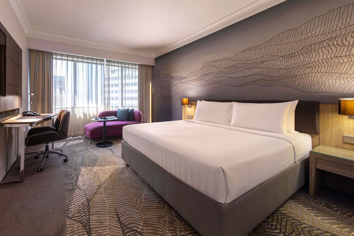 A hotel room with a large bed, a desk with a chair, a purple sofa, and a patterned carpet.