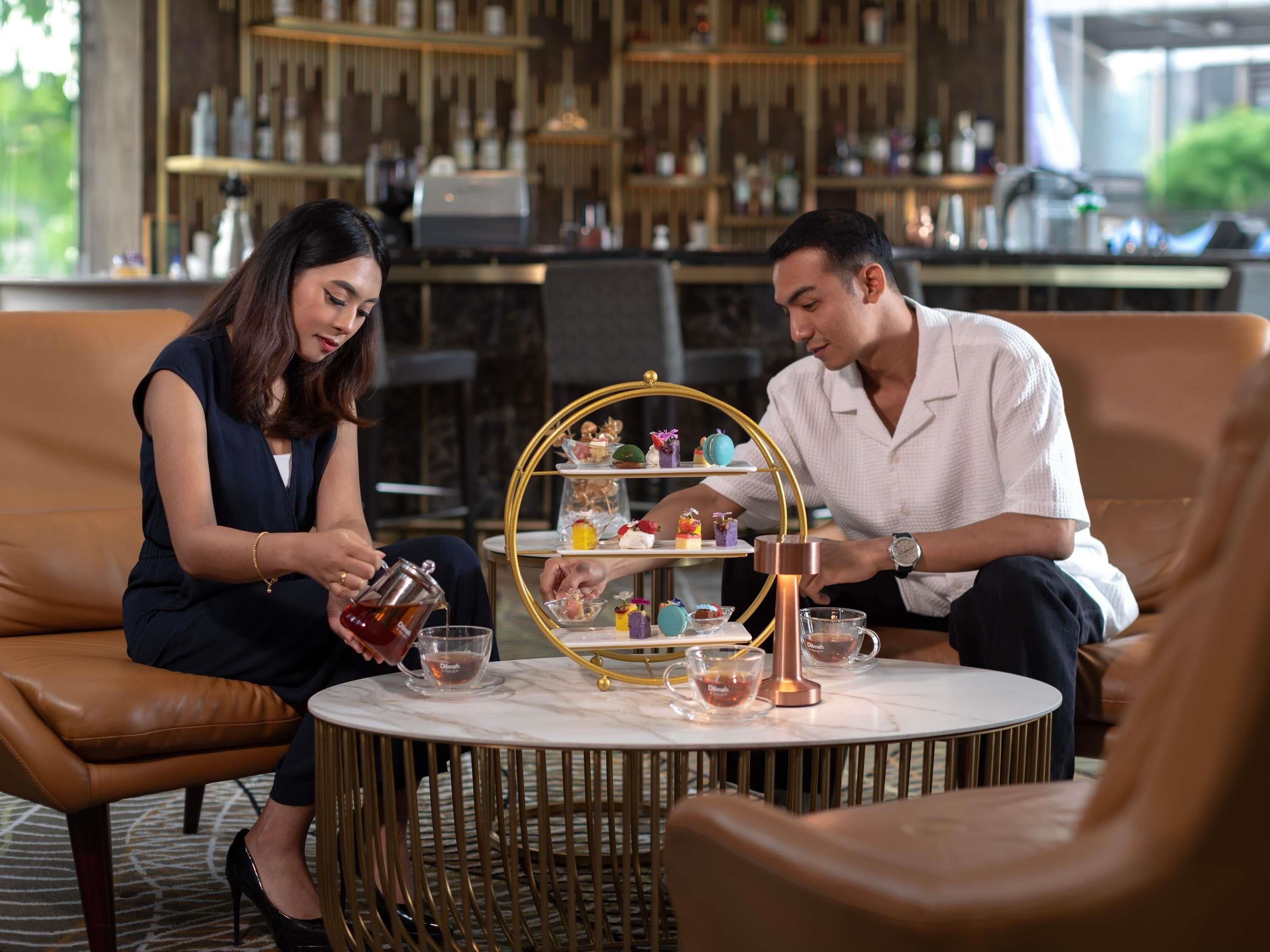 A couple seated at a round table with a tea service, surrounded by leather chairs and a decorative tray.