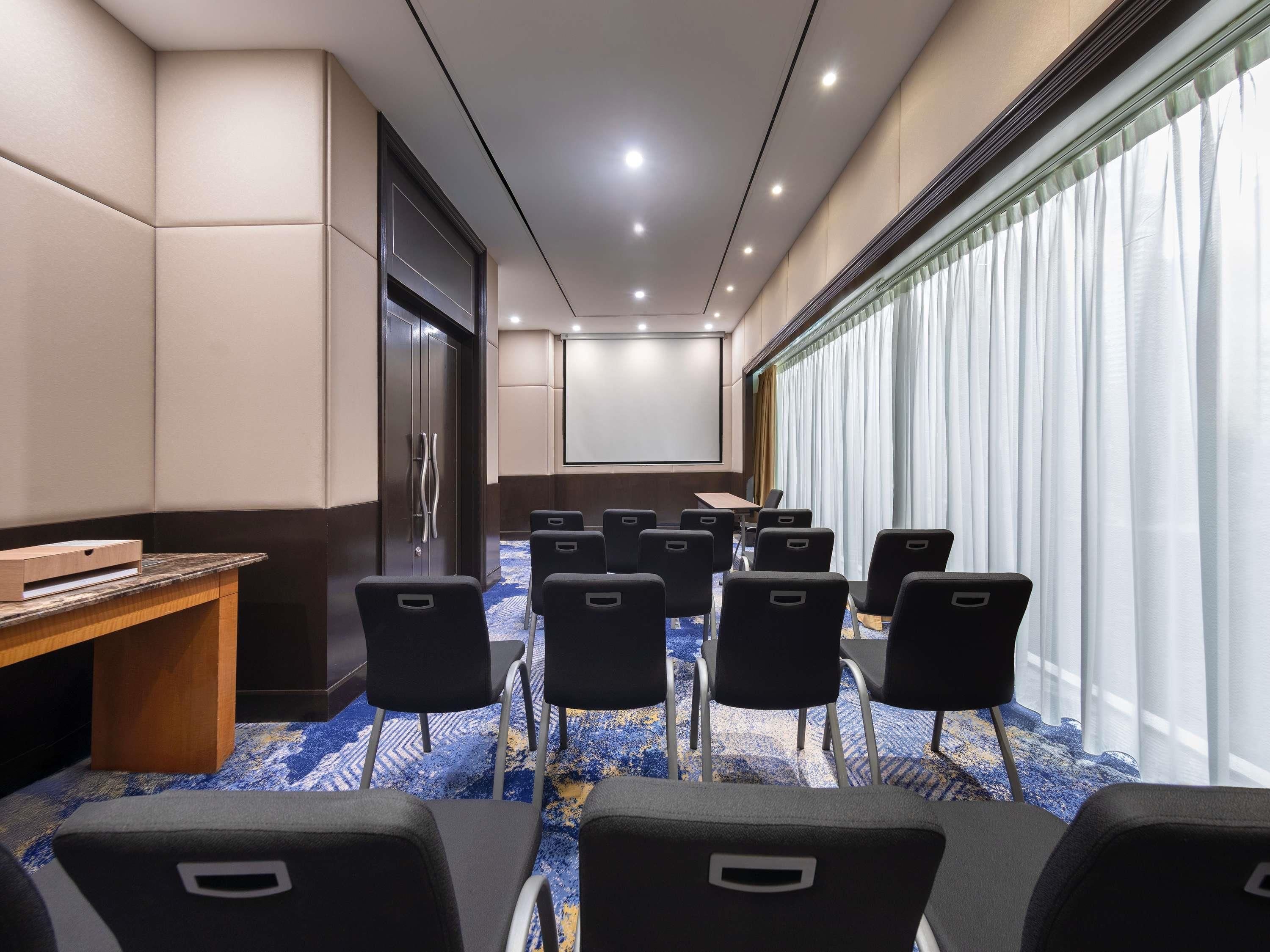 A conference room with a projector screen, a wooden desk, and rows of chairs.