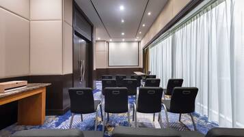 A conference room with a projector screen, a wooden desk, and rows of chairs.